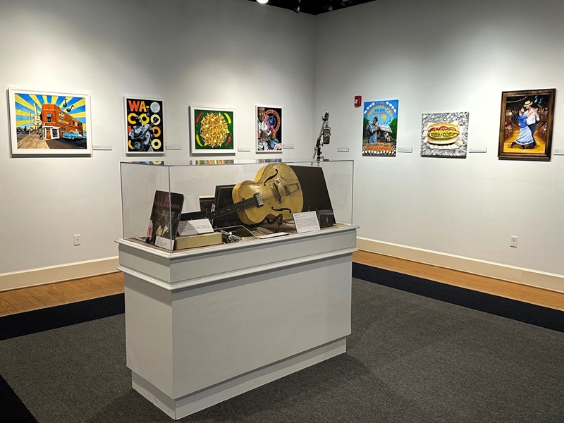 John Goodwin Exhibit Photo Three A display case with a guitar laying flat inside is centered in a room with artwork hung gallery style on the walls behind it.